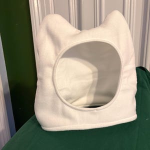 May include: A white cat-eared pet bed with a circular opening. The bed is made of soft, plush material and is designed to provide a cozy space for a cat or small pet. The bed is sitting on a green surface.