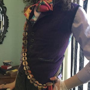 May include: A purple vest with a colorful bow tie and a long chain of spools of thread. The vest has a pocket on the front and is worn over a white shirt.