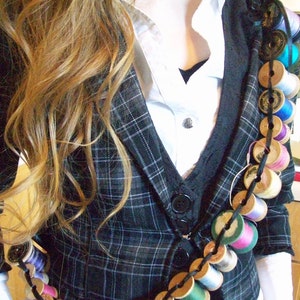 May include: A black and white plaid blazer with a white collared shirt underneath. A long chain of colorful thread spools is worn as a necklace.