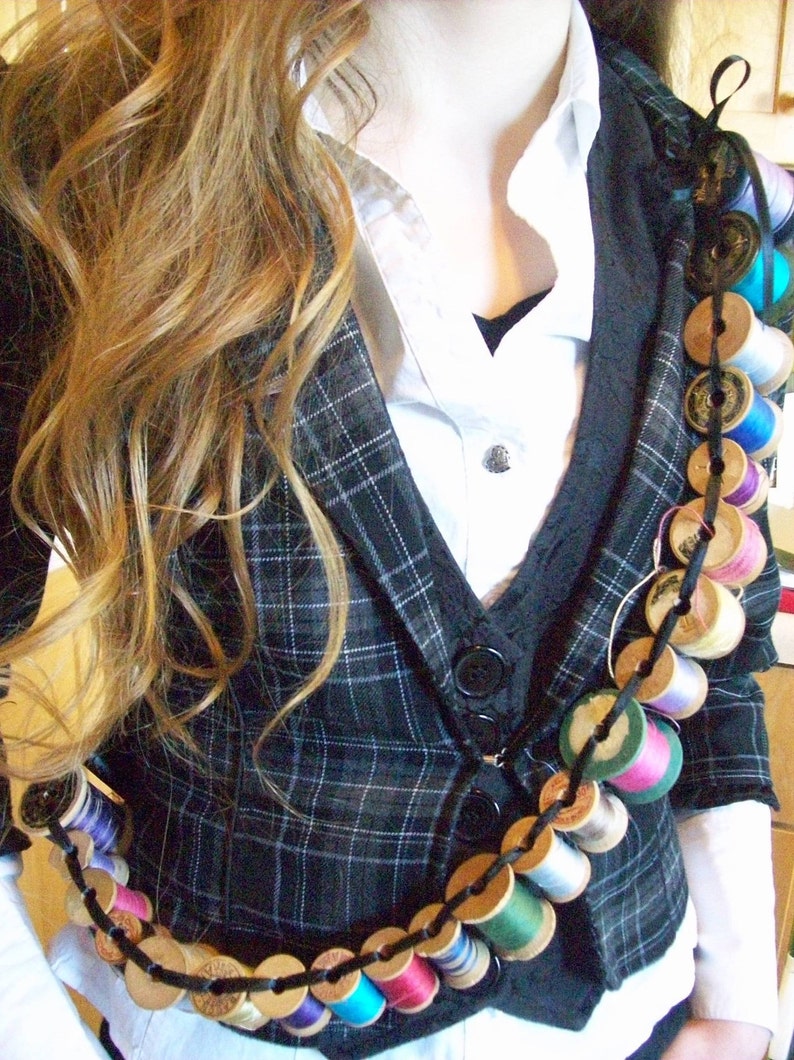 May include: A black and white plaid blazer with a white collared shirt underneath. A long chain of colorful thread spools is worn as a necklace.