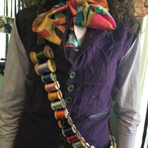 May include: A purple vest with a colorful bow tie and a long chain of spools of thread. The vest has four buttons and a pocket on the left side.