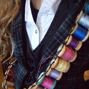 May include: A black and white plaid vest with a white collared shirt underneath. A string of colorful spools of thread is worn across the chest, attached to the vest with black string.