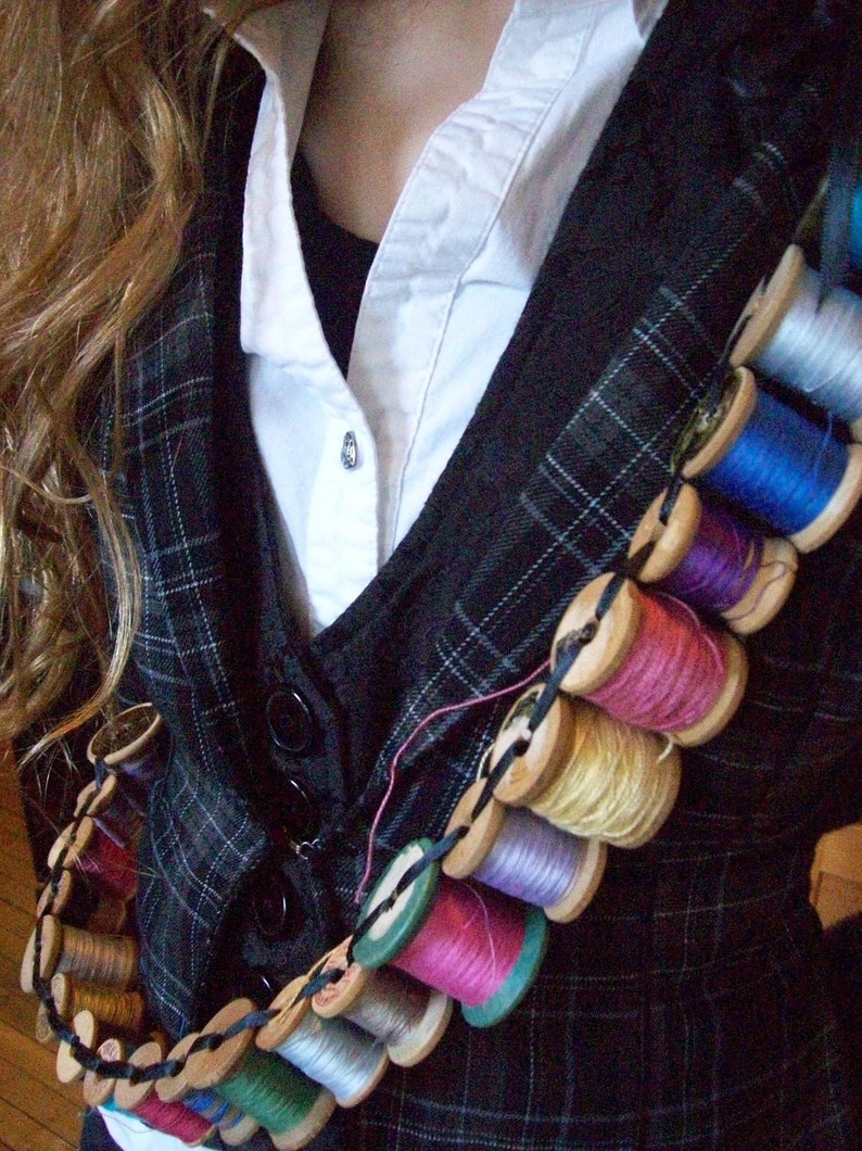 May include: A black and white plaid vest with a white collared shirt underneath. A string of colorful spools of thread is worn across the chest, attached to the vest with black string.