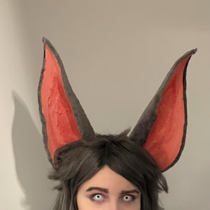 May include: Gray bat ears with a pink interior are attached to a dark gray wig. The ears are large and pointed, with a textured appearance. The person in the image has gray hair and white contact lenses.