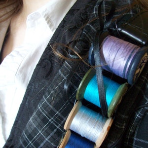 May include: A close-up of a black and white plaid jacket with four spools of thread attached to it. The spools are in various colors, including purple, blue, and white.