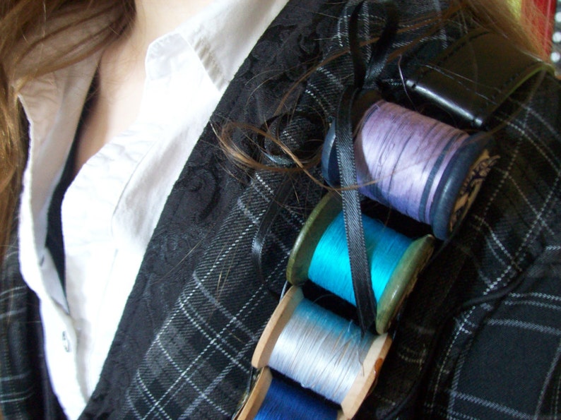 May include: A close-up of a black and white plaid jacket with four spools of thread attached to it. The spools are in various colors, including purple, blue, and white.