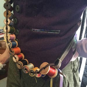 May include: A purple vest with a pocket and a long chain of wooden spools of thread wrapped around the waist. The spools are in various colors, including red, orange, yellow, green, blue, and black. The chain is attached to a brown leather strap.
