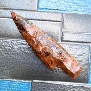 May include: A reddish-brown arrowhead with black and green inclusions. The arrowhead is pointed at one end and has a textured surface. The text "Rubycreation Store" is visible on the surface.