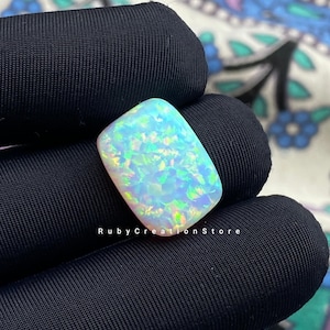 Puede incluir: Una piedra preciosa de ópalo rectangular e iridiscente con base blanca y destellos azules, verdes y amarillos. La piedra se sostiene sobre un fondo de tela negra. El texto "Ruby Creation Store" es visible.