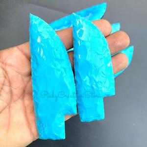 Könnte beinhalten: Zwei helltürkisfarbene, pfeilspitzenförmige Glasstücke. Das Glas hat eine strukturierte Oberfläche und ein leicht durchscheinendes Aussehen. Der Text "Ruby Creation Store" ist auf einem der Stücke sichtbar.