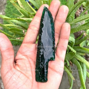 May include: A dark green, obsidian arrowhead with a textured surface. The arrowhead is held in a hand, with green plants in the background. The text "RubyCreationStore" is visible in the image.