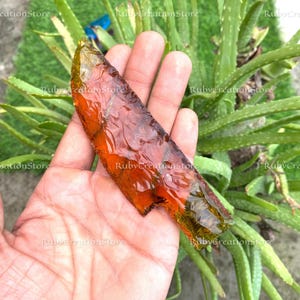May include: A translucent, amber-colored arrowhead with a textured surface. The arrowhead is held in a hand, with green plants in the background. The arrowhead has a sharp point and a jagged edge. The text "RubyCreationStore" is visible.