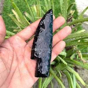 May include: A black obsidian arrowhead with a polished surface, held in a hand. The arrowhead's shape is clearly defined against a background of green foliage, highlighting its sharp edges and craftsmanship.