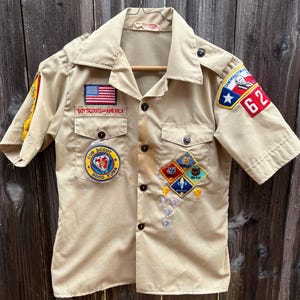 May include: Tan Boy Scouts of America uniform shirt with patches, including an American flag, the words "Boy Scouts of America", and a Cub Scout patch. The shirt has short sleeves and several other patches.