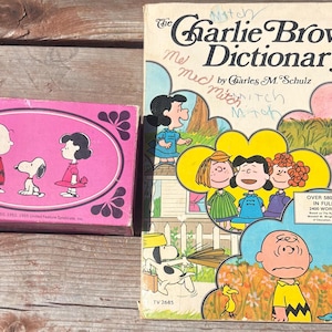 May include: Vintage children's books featuring Peanuts characters. The Charlie Brown Dictionary book is open, showing colorful illustrations. A pink box with Peanuts characters is on the left.