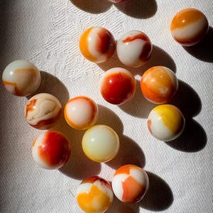 May include: A collection of fourteen spherical glass marbles, each featuring a smooth, polished surface with swirling patterns of opaque white, vibrant orange, deep red, and soft yellow colors, arranged in a scattered formation on a textured surface.