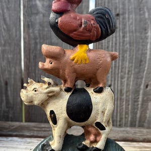 May include: A cast iron figurine featuring a stack of three farm animals on a green base. A red and black rooster stands on a light brown pig, which is balanced on a white cow with black spots. The rooster has bright yellow feet.