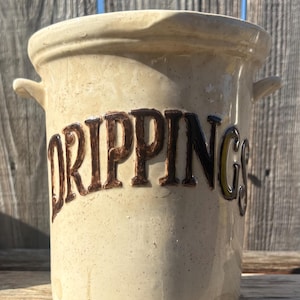 May include: A beige ceramic crock with the word "DRIPPINGS" in brown lettering. The crock has two small handles and a slightly flared rim. The background is a weathered wooden fence.
