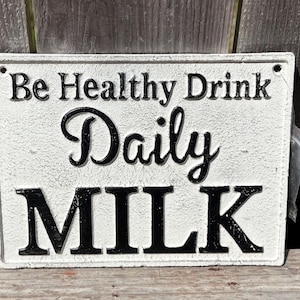 May include: Rectangular white sign with black lettering that reads "Be Healthy Drink Daily MILK". The sign has a textured surface and is mounted on a wooden background. The sign is a decorative item.