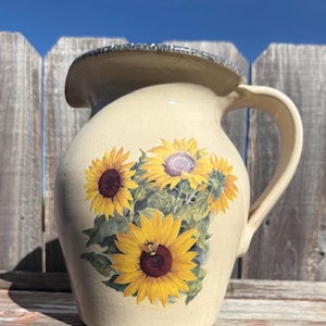 May include: Cream-colored ceramic pitcher with a handle and a spout. The pitcher is decorated with a cluster of yellow sunflowers with brown centers and green leaves. The rim has a textured, speckled design.