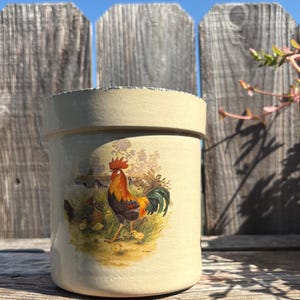 May include: A cylindrical, cream-colored ceramic jar with a detailed rooster and chick illustration. The rooster is depicted with vibrant red, orange, and green plumage. The jar has a slightly textured rim and a rustic, farmhouse aesthetic.