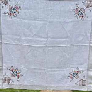 May include: A white linen tablecloth with embroidered floral designs in each corner. The embroidery features a basket-like vase filled with colorful flowers in shades of pink, blue, and yellow. The edges are finished with a simple hem.