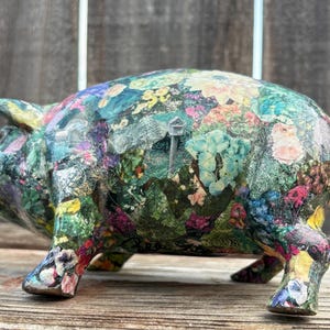 May include: A ceramic piggy bank with a colorful floral design. The pig is painted with a variety of flowers in shades of pink, blue, yellow, and green. The background is a weathered wooden surface.