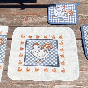 May include: A kitchen textile set featuring a white towel, a square washcloth, a square pot holder, and an oven mitt. Each item displays a blue and white checkered pattern with a white rooster illustration and small orange chick accents.