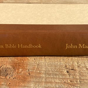May include: The MacArthur Bible Handbook by John MacArthur, a brown leather-bound book with gold lettering.