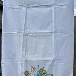May include: White linen towel with an embroidered church scene. The design features a church with a blue steeple, green trees, and pink flowers. The bottom edge has a green crocheted trim.