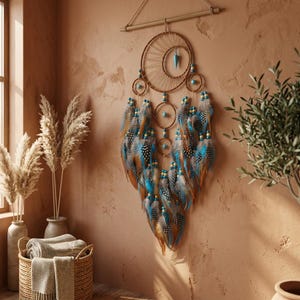 Op de afbeelding: Een bruine dromenvanger met een grote centrale hoepel en kleinere hoepels eronder, versierd met blauwe en bruine veren. De dromenvanger hangt aan een houten deuvel en heeft turquoise kralen. Decoratieve wandkunst.