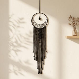 Op de afbeelding: Een zwarte macramé wandhanger met een cirkelvormig frame, een geweven webpatroon, een centrale donkere kraal en lange, trapsgewijze zwarte koordfranjes met knoopdetails en kleine kwastjes aan de uiteinden die verticaal naar beneden hangen.