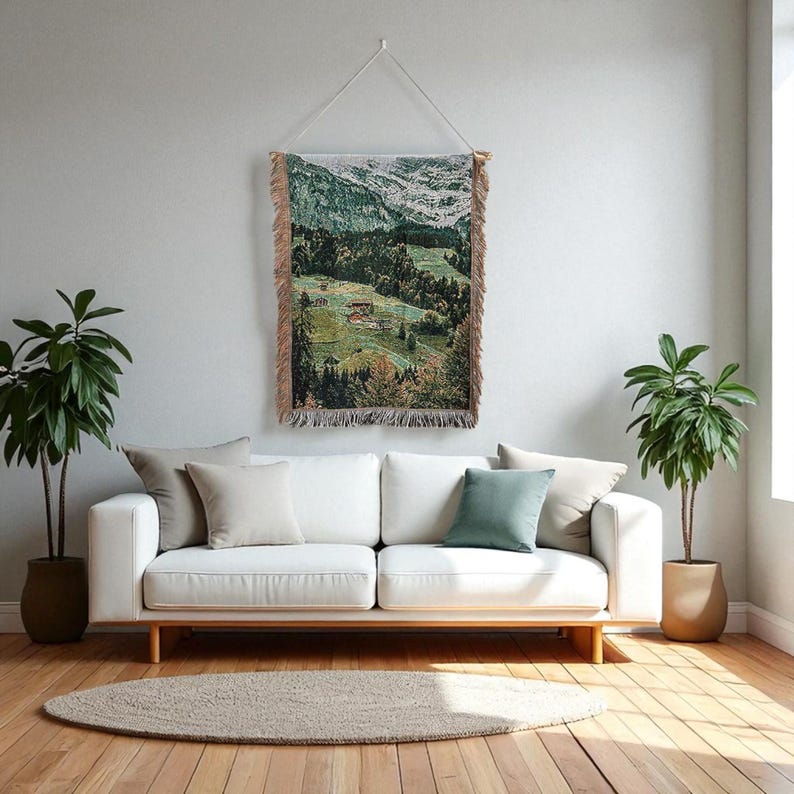 May include: A woven wall hanging featuring a mountain scene with green forests, snowy peaks, and buildings. The tapestry has a fringed edge and is hung from a wooden rod. The colour scheme includes greens, browns, and whites.