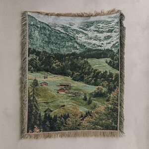 May include: Woven throw blanket depicting a mountain landscape with green hills and small buildings. The blanket, with a fringed edge, is predominantly green, brown, and white. The design creates a sense of nature and peace, perfect for a cosy home.