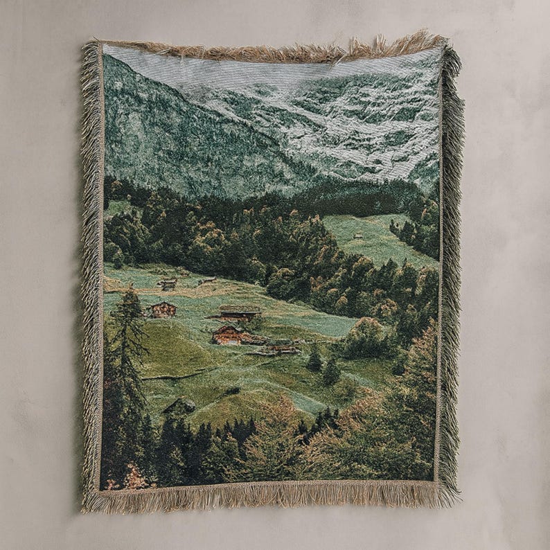 May include: Woven throw blanket depicting a mountain landscape with green hills and small buildings. The blanket, with a fringed edge, is predominantly green, brown, and white. The design creates a sense of nature and peace, perfect for a cosy home.