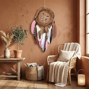 May include: A handcrafted dreamcatcher with a woven circular frame, adorned with pink, white, and brown feathers. The dreamcatcher hangs against a warm, earthy-toned wall, creating a bohemian aesthetic. The feathers are approximately 10 inches long.