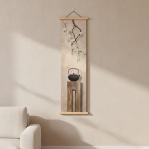 Puede incluir: Un tapiz vertical con un fondo beige, una rama de flores blancas y una tetera negra sobre un soporte de madera. La obra de arte está enmarcada con madera clara y suspendida por una cuerda.