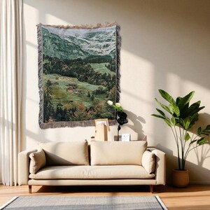 May include: A rectangular woven tapestry featuring a mountain scene with green forests, snowy peaks, and small buildings. The tapestry has a fringed border and is displayed on a beige wall above a beige sofa.