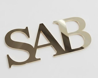 May include: Gold-coloured metal letters spelling out the word "SAB". The letters are in a sans-serif font and are slightly angled, casting subtle shadows. The background is plain white.