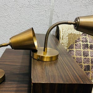 May include: Two brass-coloured metal lamps with round bases and flexible gooseneck arms. Each lamp features a conical shade. The lamps have a metallic finish and are positioned on a dark wood surface, showcasing their adjustable design.