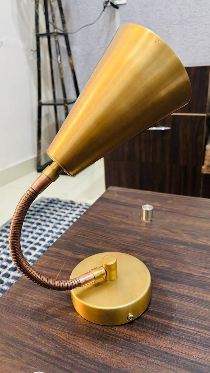 May include: A gold-toned metal desk lamp featuring a conical shade, a flexible copper-coloured gooseneck arm, and a circular base. The lamp has a smooth, matte finish and a simple, functional design with a single screw visible on the side of the base.