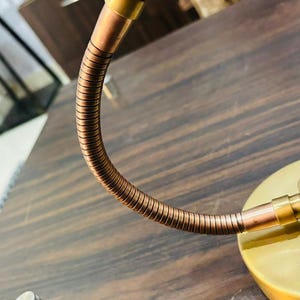 May include: A desk lamp featuring a smooth, matte gold-toned conical shade and a circular base. The lamp includes a flexible, ribbed copper-coloured gooseneck arm that connects the shade to the base, allowing for adjustable positioning of the light.