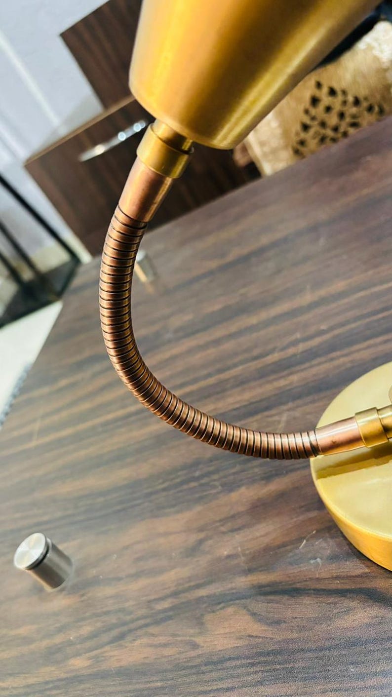 May include: A desk lamp featuring a smooth, matte gold-toned conical shade and a circular base. The lamp includes a flexible, ribbed copper-coloured gooseneck arm that connects the shade to the base, allowing for adjustable positioning of the light.