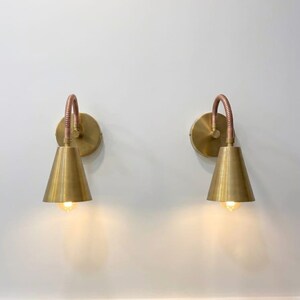 May include: Two wall-mounted lamps featuring circular brass backplates, flexible copper-toned gooseneck arms, and conical brass shades. Each lamp emits a warm glow from the base of the shade, providing focused downward illumination.