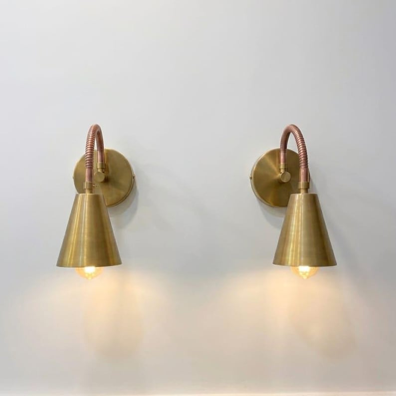 May include: Two wall-mounted lamps featuring circular brass backplates, flexible copper-toned gooseneck arms, and conical brass shades. Each lamp emits a warm glow from the base of the shade, providing focused downward illumination.