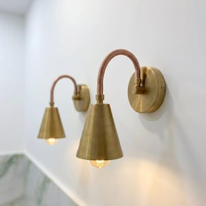 May include: Two wall-mounted light fittings featuring circular brass backplates, flexible copper-toned gooseneck arms, and conical brass shades. Each fitting holds a single light bulb that emits a warm glow, creating a focused downward illumination.