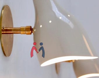 May include: A cream-coloured wall-mounted light fitting with brass detailing. The fixture features a curved shade and a circular base. The light is switched on, casting a glow downwards. The design is modern and minimalist.