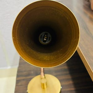 May include: A brass-toned metal desk lamp featuring a conical shade viewed from directly above, revealing a central light bulb socket. The lamp has a circular base connected to the shade by a short, adjustable arm, all in a matching metallic finish.
