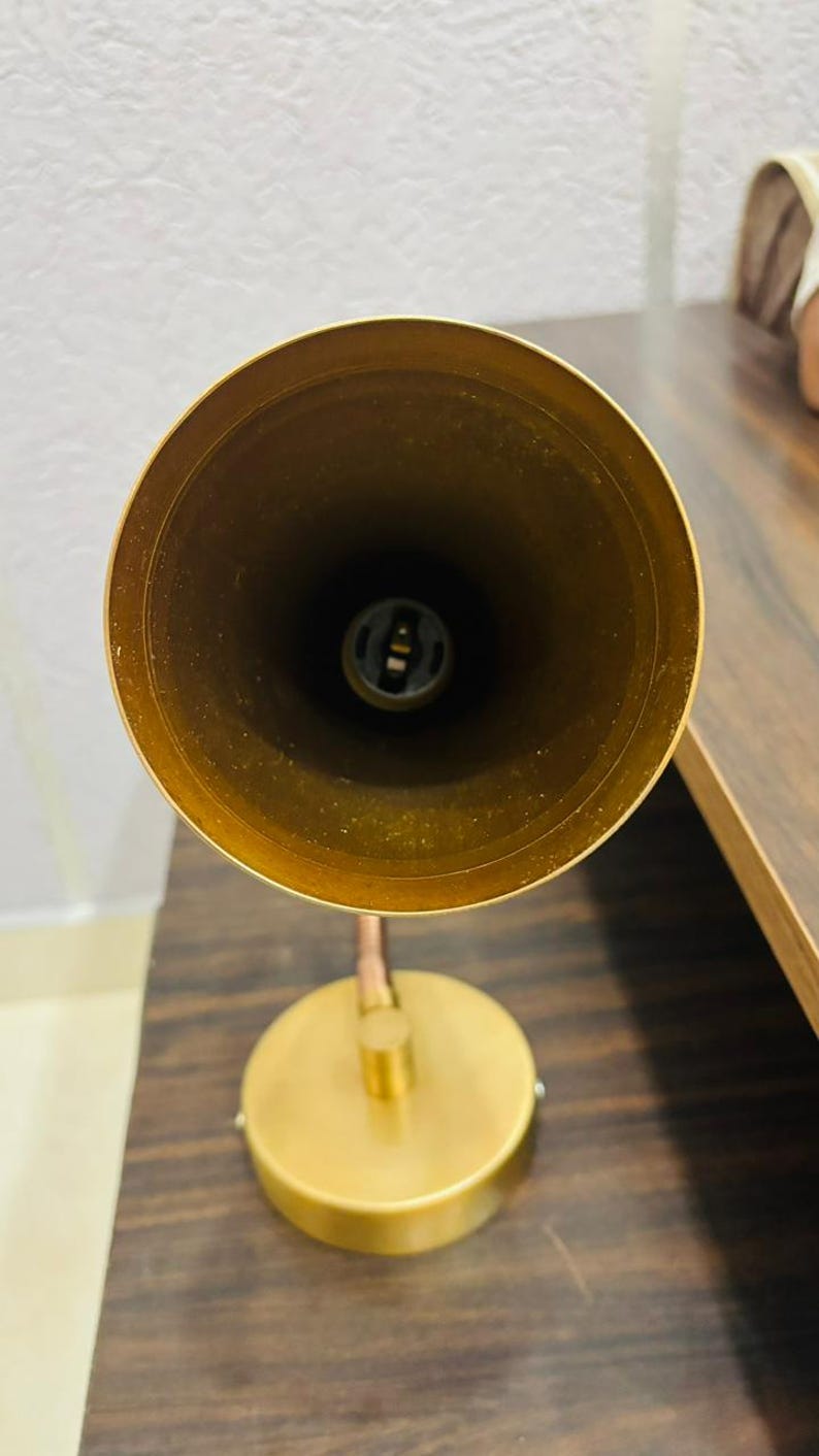 May include: A brass-toned metal desk lamp featuring a conical shade viewed from directly above, revealing a central light bulb socket. The lamp has a circular base connected to the shade by a short, adjustable arm, all in a matching metallic finish.
