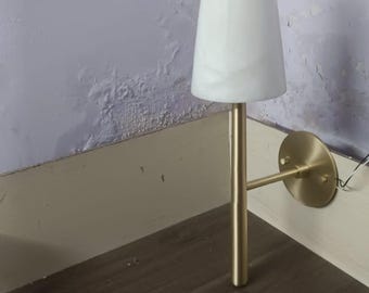 May include: A wall-mounted sconce light fitting featuring a brushed gold metal arm and a circular wall plate. The lamp includes a conical, white, semi-translucent shade with subtle marbled veining, positioned vertically on the slender metal stem.